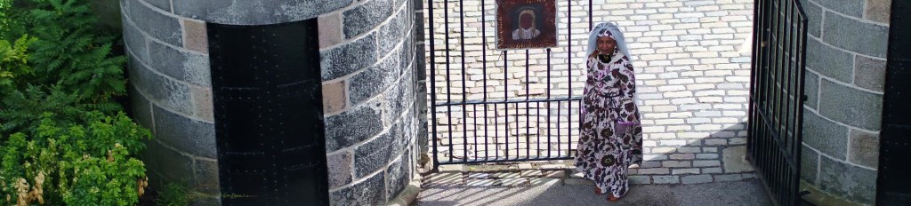 Looking down on round granite tower black iron gate and a black woman dressed in floor length patterned gown and veil looks up. Shadows slant from the gate over the cobbles.