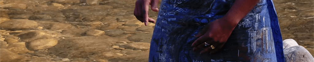 detail of black woman's hands wearing blue paper skirt with drawings on with background of clear sunlit river flowing over pebbles